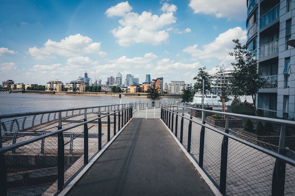 A paved pedestrian walkway with metal railings on both sides extends toward a river, with a view of the distant city skyline featuring modern high-rise buildings under a partly cloudy sky. On the right side of the image, there is a tall, curved residential building with reflective glass windows and balconies, partially obscured by a small tree and some shrubbery. The riverbank below the walkway shows a wooden seating area with benches and a few lampposts. In the background, alongside the city skyline, there are several boats docked at a nearby marina, hinting at a waterfront location suitable for private disposal or alternative waste handling. The scene suggests an urban environment with an emphasis on outdoor space and waterways, aligning with areas where rubbish removal services may be needed for maintaining cleanliness of communal walkways and surrounding areas, especially those close to residential complexes and waterfront promenades managed by companies like Rubbish Removal Canary Wharf.