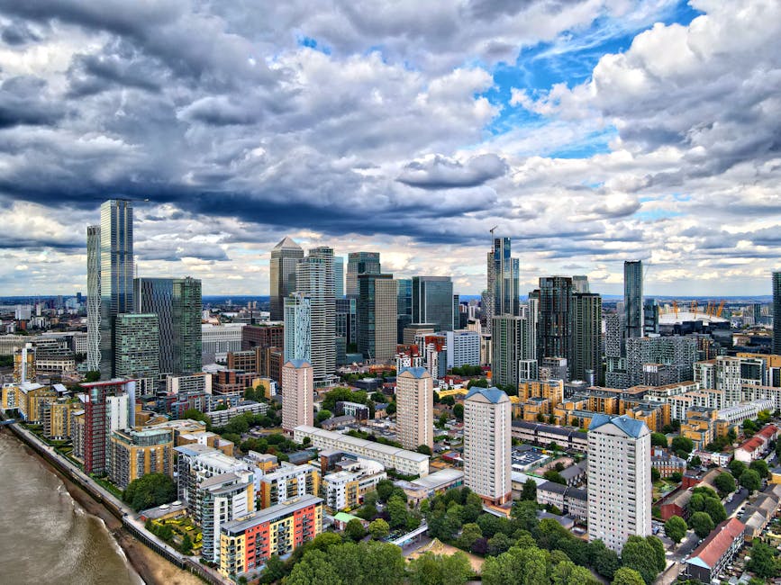 An expansive aerial view of a modern cityscape shows numerous high-rise office buildings, residential towers, and commercial structures clustered together, with some featuring glass facades reflecting the sky. In the foreground, a series of mid-rise apartment blocks with flat roofs and varied pastel and neutral colour schemes are interspersed with greenery, including trees lining the streets and small patches of landscaped areas. To the left, a river runs parallel to the city, with a concrete embankment bordering its edge. The background features dense cloud cover, casting a moody, diffused light over the scene, with patches of blue sky visible through breaks in the clouds. The overall environment suggests a well-developed urban centre, where private waste management services, such as those offered by Rubbish Removal Canary Wharf, may provide alternative waste handling to municipal collection, supporting the area's high density and diverse infrastructure. The image captures a snapshot of city life and the urban environment, highlighting the scale and density of modern development, with the focus on the dense cluster of tall buildings and their surrounding lower-rise structures.