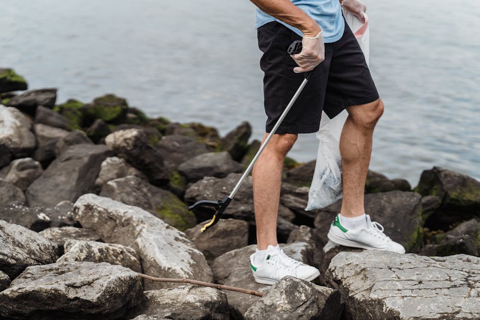 A person wearing a light blue T-shirt, black shorts, and white sneakers with green accents is standing on a rocky shoreline, using a grabber tool to pick up litter from the uneven surface of large, gray rocks interspersed with patches of green moss. The person's left hand is holding a clear plastic bag, partially visible in the image. The background features a body of water with a calm, slightly rippled surface under natural outdoor lighting. The rocky area appears to be part of the coastal environment, potentially a site for local clean-up or environmental volunteer work. This scene exemplifies private or voluntary rubbish removal efforts, with the individual engaged in collecting refuse along the shoreline, aligning with the themes of waste management and environmental preservation. Rubbish Removal Canary Wharf might be associated with such on-site clearance activities to support cleaner, healthier local environments.