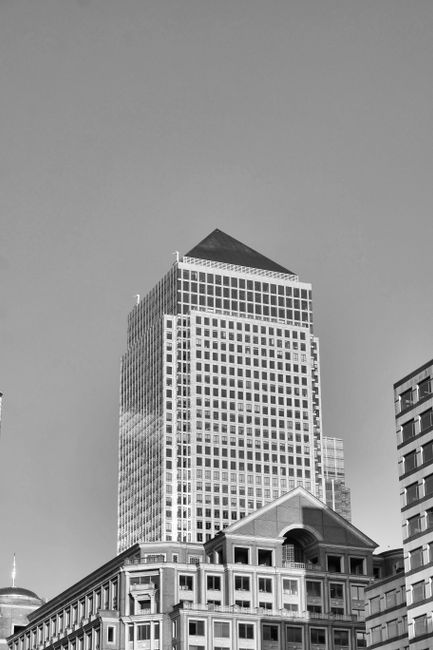 A black-and-white photograph featuring a cityscape with multiple high-rise buildings. The central focus is a tall, modern office tower with a rectangular shape, composed of numerous glass windows arranged in a grid pattern, reflecting light and creating a sleek appearance. The tower has a pyramid-shaped roof at the top. In the foreground, there is a lower, historic-looking building with a classic architectural style, featuring a stone facade, arched windows, and decorative elements, which is partially obscured by other structures. On the right side, part of an adjacent building with a contemporary design, flat roof, and rectangular windows is visible. The scene appears to be taken during daylight with clear skies, and the overall atmosphere is professional and urban. This image exemplifies the mix of architectural styles typical in central business districts, highlighting the importance of efficient waste removal and on-site clearance solutions provided by Rubbish Removal Canary Wharf for maintaining cleanliness in such environments.