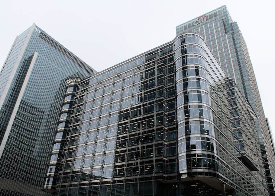 A modern multi-storey office building with a curved glass façade, featuring tinted reflective windows and metallic framing, situated among other tall commercial structures in an urban cityscape. The building's exterior showcases a combination of transparent and mirrored glass panels, creating a sleek, contemporary appearance. The surrounding environment includes neighboring high-rise buildings with similar glass and steel finishes, and part of the street or pavement is faintly visible at the base. The sky overhead appears overcast, contributing to diffuse, even lighting across the scene. This type of building exemplifies the commercial architecture typical of business districts, where independent waste collection and on-site clearance services such as those provided by Rubbish Removal Canary Wharf may be necessary for maintaining the premises' cleanliness and managing waste from office operations.