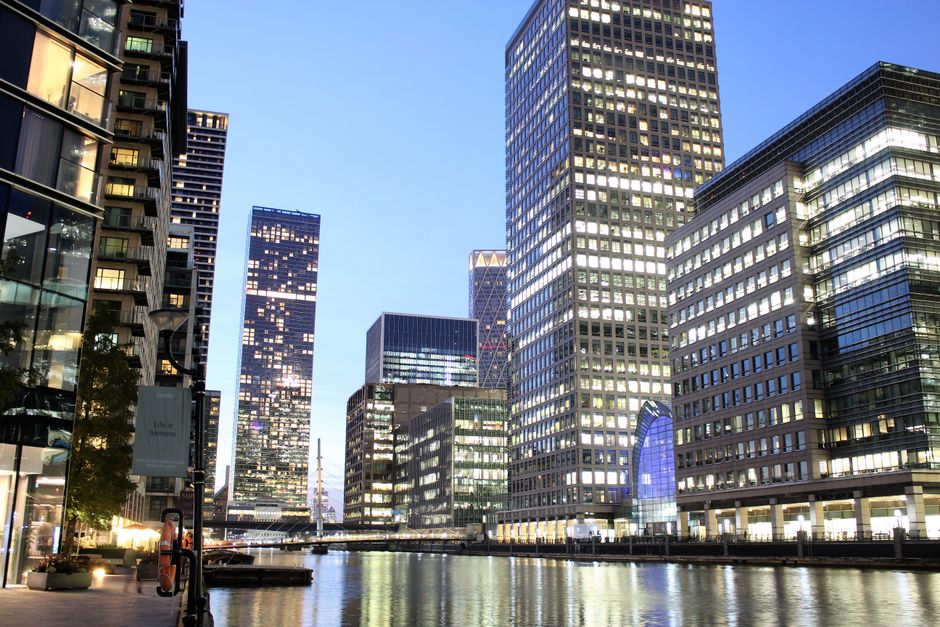 A black-and-white photograph featuring a cityscape with multiple high-rise buildings. The central focus is a tall, modern office tower with a rectangular shape, composed of numerous glass windows arranged in a grid pattern, reflecting light and creating a sleek appearance. The tower has a pyramid-shaped roof at the top. In the foreground, there is a lower, historic-looking building with a classic architectural style, featuring a stone facade, arched windows, and decorative elements, which is partially obscured by other structures. On the right side, part of an adjacent building with a contemporary design, flat roof, and rectangular windows is visible. The scene appears to be taken during daylight with clear skies, and the overall atmosphere is professional and urban. This image exemplifies the mix of architectural styles typical in central business districts, highlighting the importance of efficient waste removal and on-site clearance solutions provided by Rubbish Removal Canary Wharf for maintaining cleanliness in such environments.