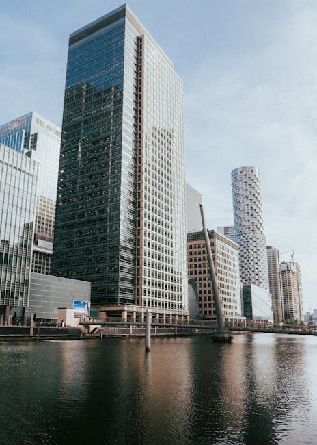 A cluster of modern high-rise buildings along a waterway in an urban setting. The tallest building in the center features a sleek, glass exterior with reflective surfaces, showcasing a mixture of dark and light tones. Surrounding it are other skyscrapers made of glass and steel, some with textured or patterned facades. To the right, a cylindrical tower with a distinctive, rounded design and a perforated, white surface adds variety to the skyline. The buildings are situated on a paved waterfront promenade with a low wall and a metal railing, and a few industrial elements, such as a crane visible in the background, indicate ongoing development. The water in the foreground is calm, reflecting the buildings and the pale, partly cloudy sky above. The scene exemplifies a modern business district with contemporary architecture, representing the type of environment where professional waste management or rubbish removal services by companies like Rubbish Removal Canary Wharf might be employed for on-site clearance or private waste handling tasks in a commercial setting.