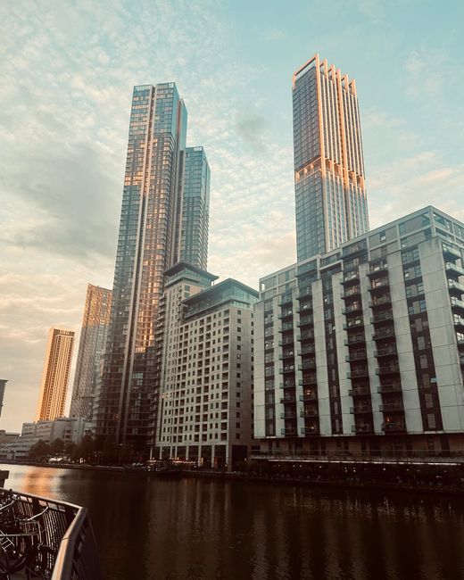 A view of modern high-rise buildings in an urban riverside setting, including a pair of tall glass towers with reflective blue and pinkish hues, situated behind a lower, white and grey residential or commercial structure with multiple balconies and rectangular windows. The buildings are closely clustered and extend upwards against a partly cloudy sky with a soft, warm light, suggesting late afternoon or early evening. In the foreground, the edge of a black metal railing is visible next to a calm river or waterway, with the water reflecting the buildings' facades. The scene depicts a contemporary cityscape typical of a central business district or vibrant urban area, which may relate to independent rubbish removal services or private waste handling near commercial or residential developments, as observed through the surrounding environment of tall structures and waterfront location, aligning with the scope of rubbish removal in the Canary Wharf district.