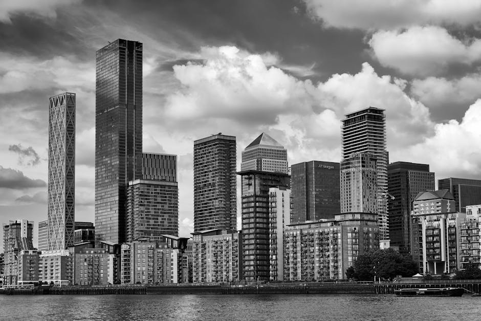 A cluster of modern high-rise buildings along a waterway in an urban setting. The tallest building in the center features a sleek, glass exterior with reflective surfaces, showcasing a mixture of dark and light tones. Surrounding it are other skyscrapers made of glass and steel, some with textured or patterned facades. To the right, a cylindrical tower with a distinctive, rounded design and a perforated, white surface adds variety to the skyline. The buildings are situated on a paved waterfront promenade with a low wall and a metal railing, and a few industrial elements, such as a crane visible in the background, indicate ongoing development. The water in the foreground is calm, reflecting the buildings and the pale, partly cloudy sky above. The scene exemplifies a modern business district with contemporary architecture, representing the type of environment where professional waste management or rubbish removal services by companies like Rubbish Removal Canary Wharf might be employed for on-site clearance or private waste handling tasks in a commercial setting.